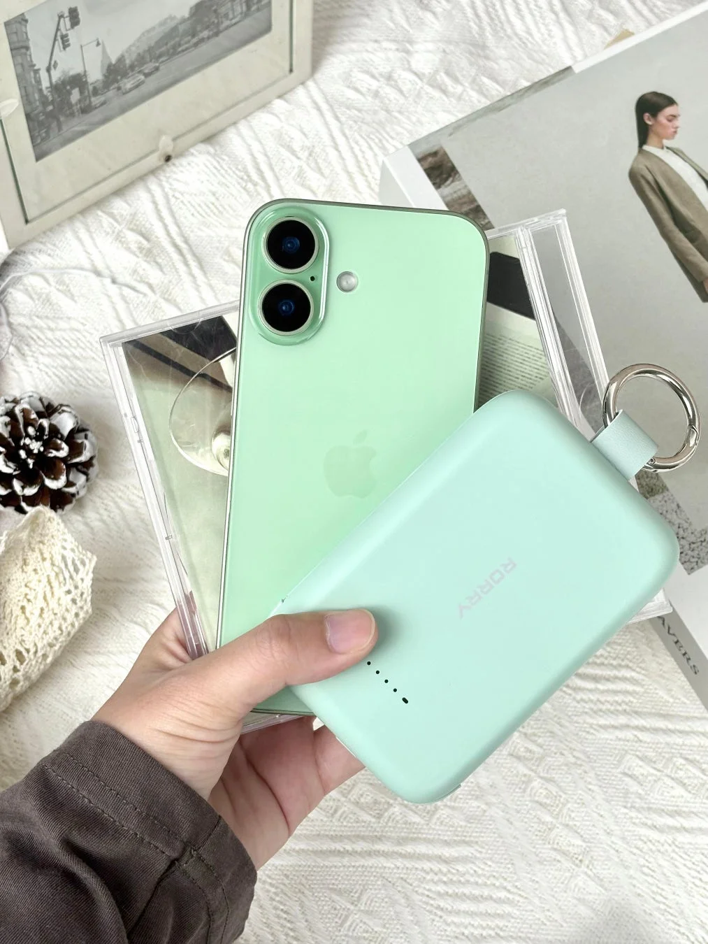 ミントグリーンのiPhoneと、同じ色のコンパクトなモバイルバッテリーを手に持っている様子が写っています。背景には白地の布や写真立て、雑誌が配置されており、デジタル製品と日常が融合したイメージです。