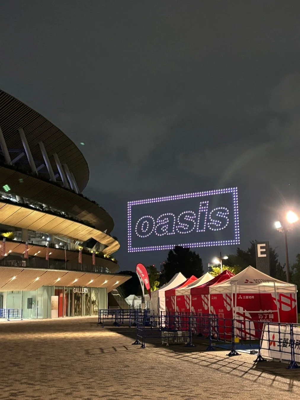 夜のスタジアムを背景に、「oasis」の文字がドローンライトショーで大きく浮かび上がっています。手前にはイベント用の赤いテントや入場受付の案内が見られ、大規模な屋外イベントが開催されている様子がうかがえます。