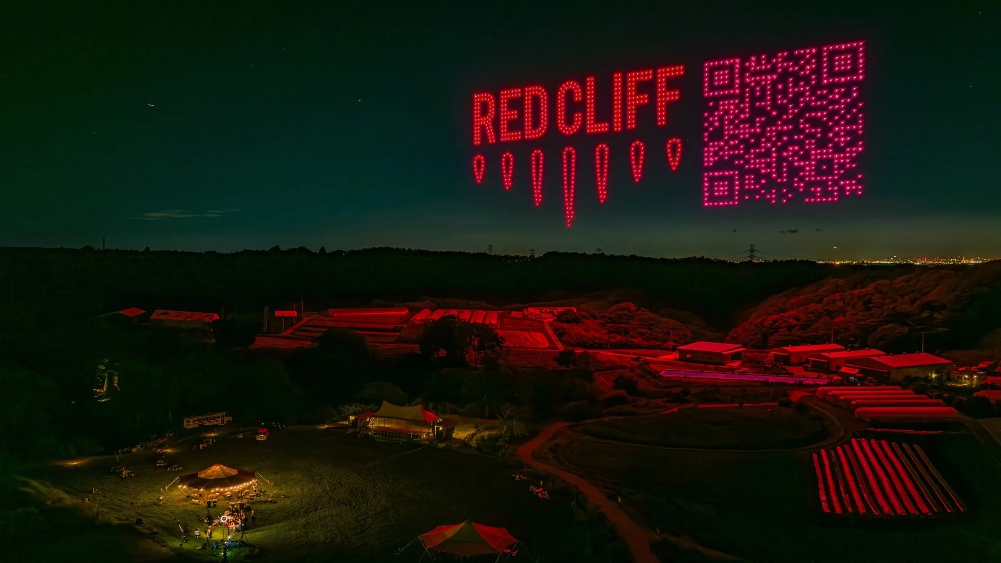 夜空に「RED CLIFF」の文字とQRコードがドローンで描かれ、地上では赤い照明が印象的な屋外施設や畑が広がっています。イベントが開催されているようです。