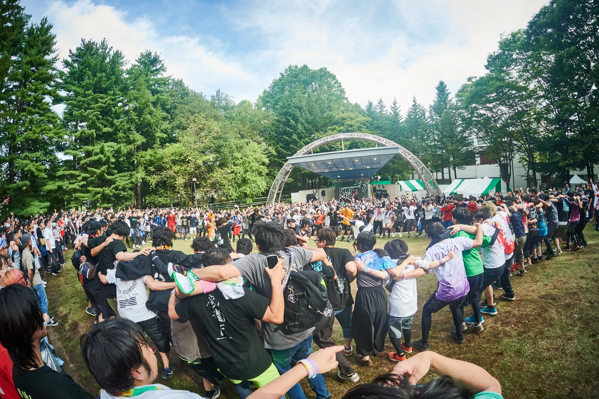 青空の下、野外音楽フェスで多くの観客がステージ前に集まり、肩を組み大きな輪になって盛り上がっている