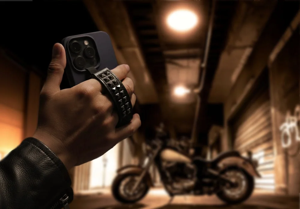 ライダースジャケットを着た人物が、スタッズ付きのストラップが付いたスマートフォンを持っている。背景にはぼやけたバイクと暗い路地が広がる、クールな雰囲気の画像です。