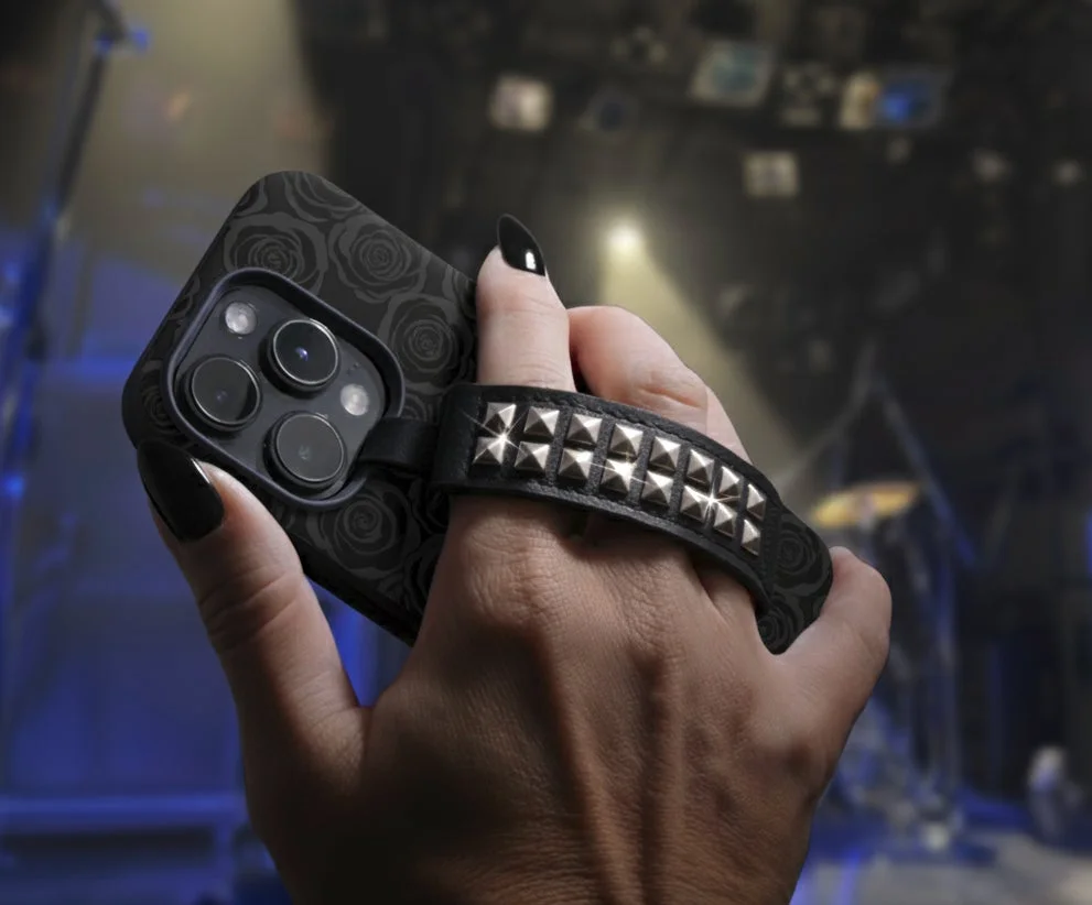 黒いネイルを施した手が、黒いバラ柄のスマホケースとスタッズ付きストラップが付いたスマートフォンを持っているクローズアップ。背景にはライブ会場のような照明が見え、ゴシックやロックテイストのファッションを想起させる。