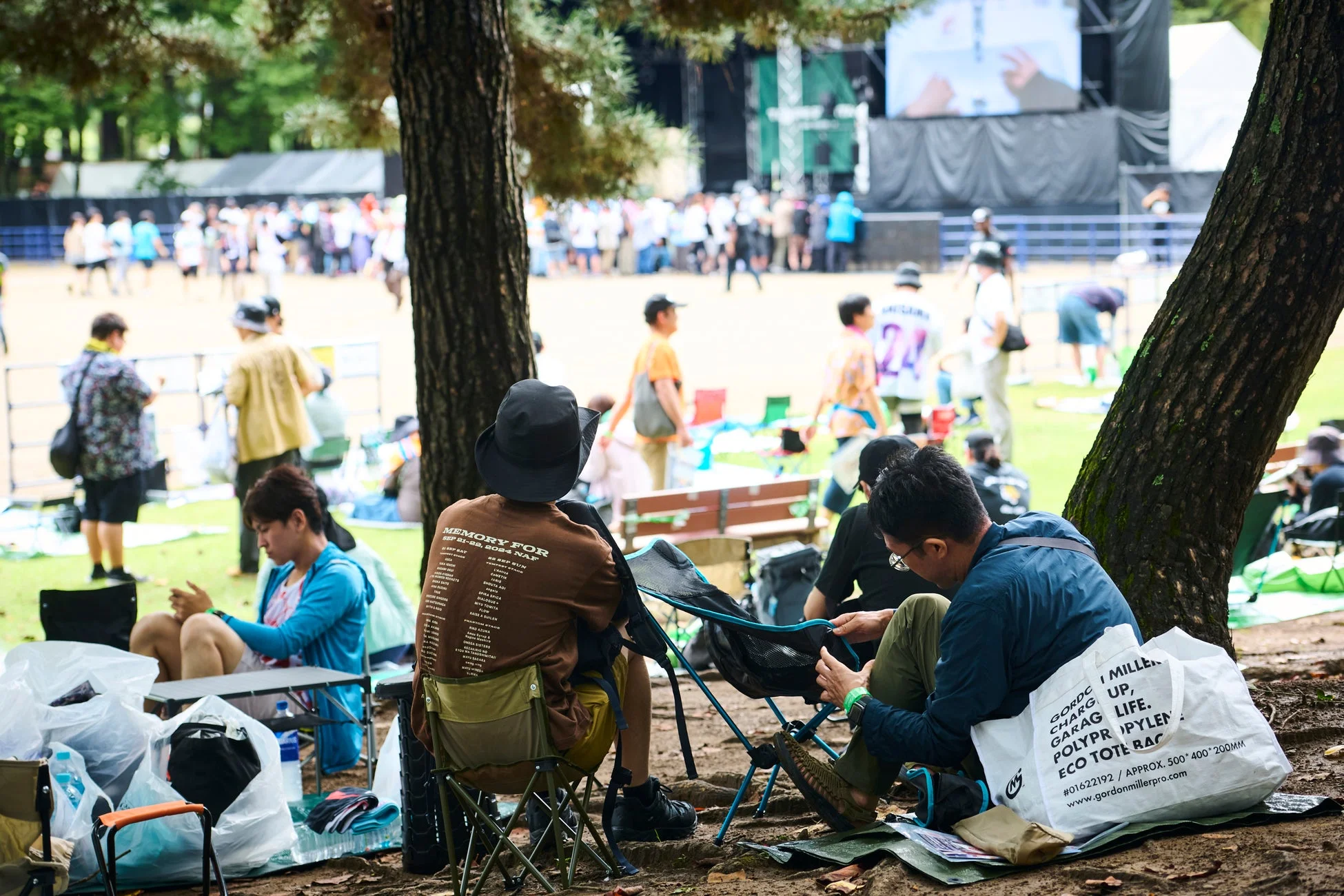 公園の木陰でくつろぐ人々が写っています。背景にはステージが見え、屋外イベントやフェスティバルを楽しんでいる様子です。