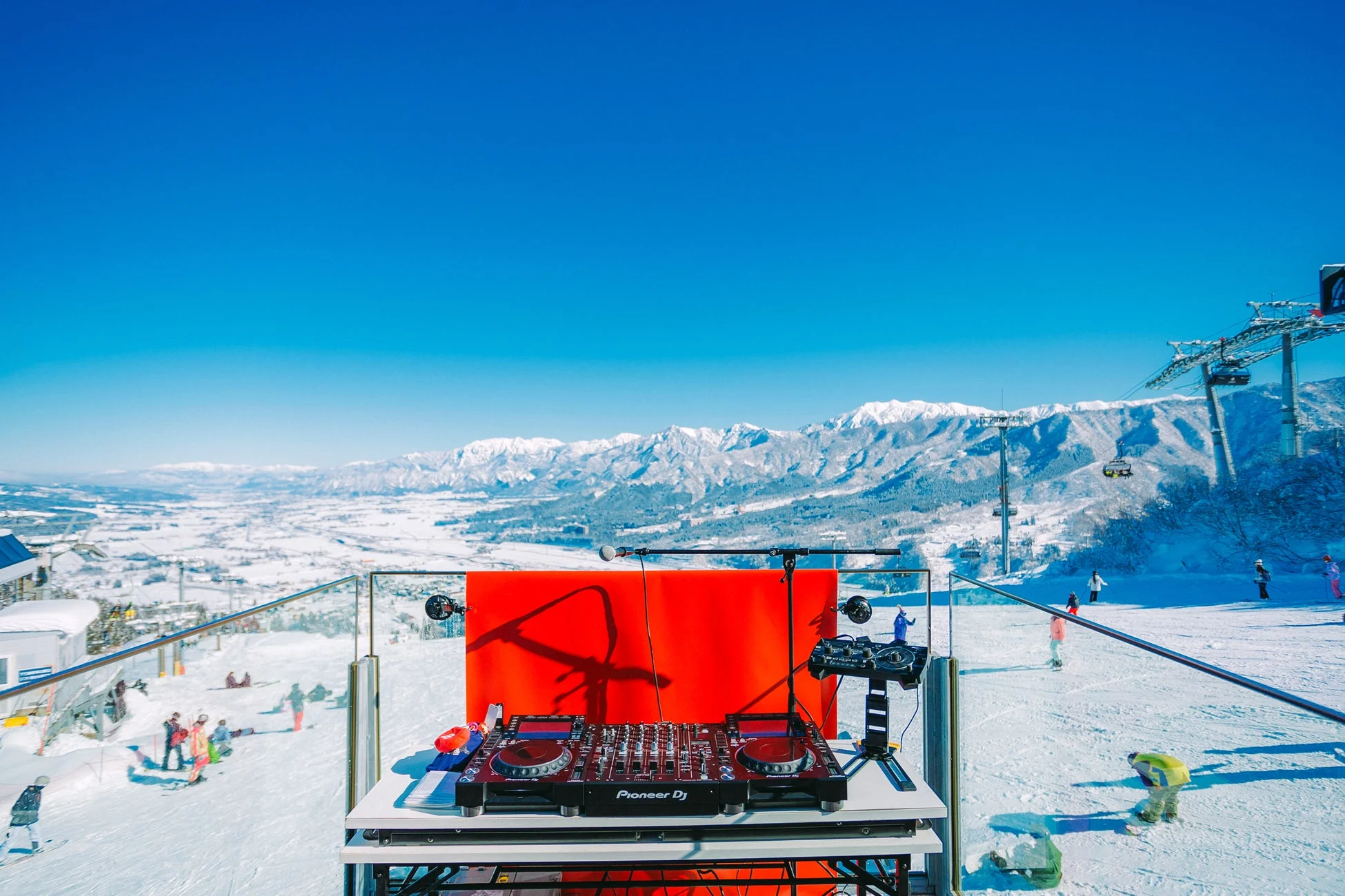 晴れた冬のスキー場で、DJブースが設置され、壮大な雪山を背景に音楽イベントが開催されている様子です。ゲレンデではスキーやスノーボードを楽しむ人々が見られ、活気あふれる冬の風景が広がっています。