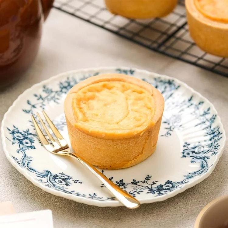 美しい模様の皿に置かれた、焼き色のついたチーズタルトとゴールドのフォーク。奥にはティーポットが見え、優雅なティータイムを思わせる一枚です。