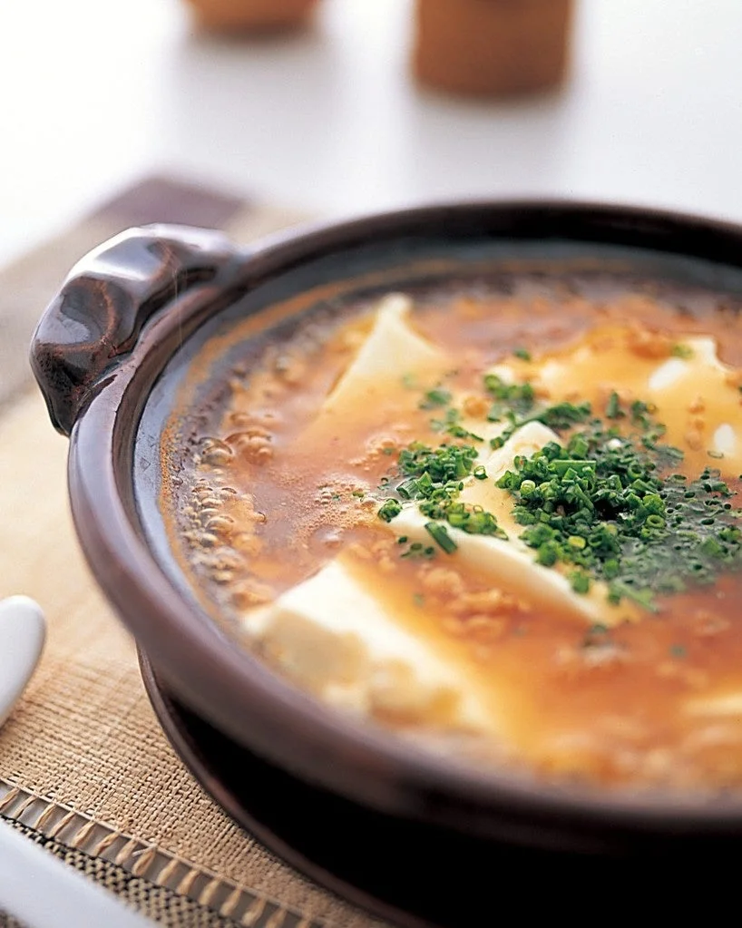 土鍋に入った温かい豆腐料理のクローズアップ。白い豆腐が茶色の出汁に浸かり、細かく刻まれた青ネギが薬味として添えられています。湯気が立ち上るような温かみがあり、ヘルシーな和食の雰囲気を伝える一枚です。