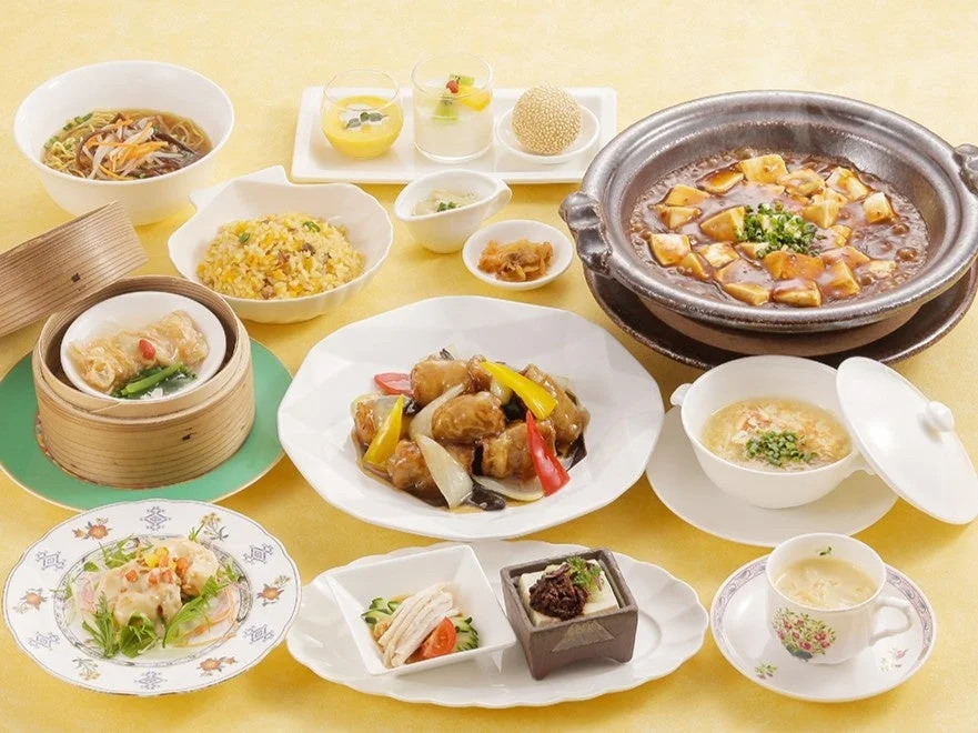 黄色のテーブルクロスに並べられた豪華な中華料理のフルコースセット。麻婆豆腐、酢豚、炒飯、ラーメン、点心、サラダ、デザートなど、様々な料理が豊富に盛り付けられており、見た目にも華やかで食欲をそそる。