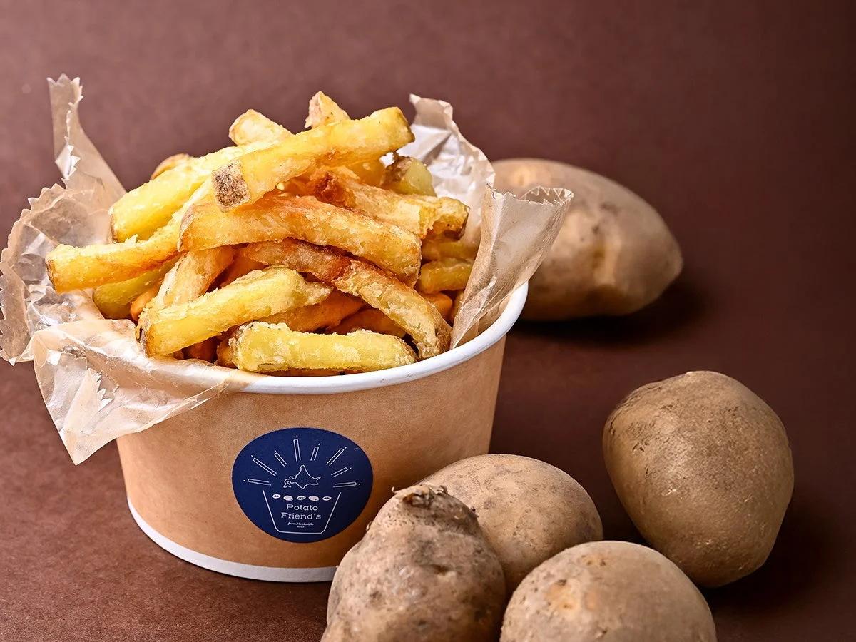 揚げたてのフライドポテトが紙カップに盛られ、周囲に生のじゃがいもが複数置かれています