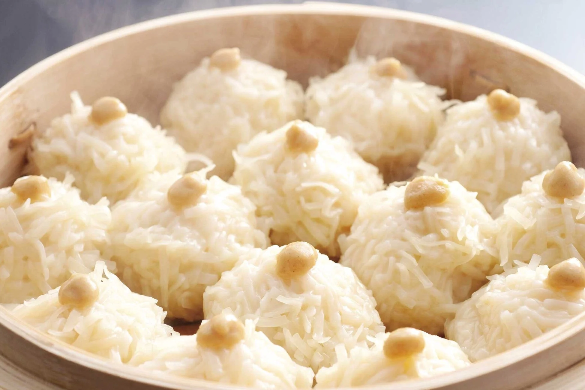 竹製の蒸し器に盛られた、温かい中華点心の写真です。白米で包まれた丸い焼売のような料理が多数並び、それぞれの上に小さなトッピングが乗っています。湯気が立ち上っており、出来立ての美味しそうな様子が伝わります。