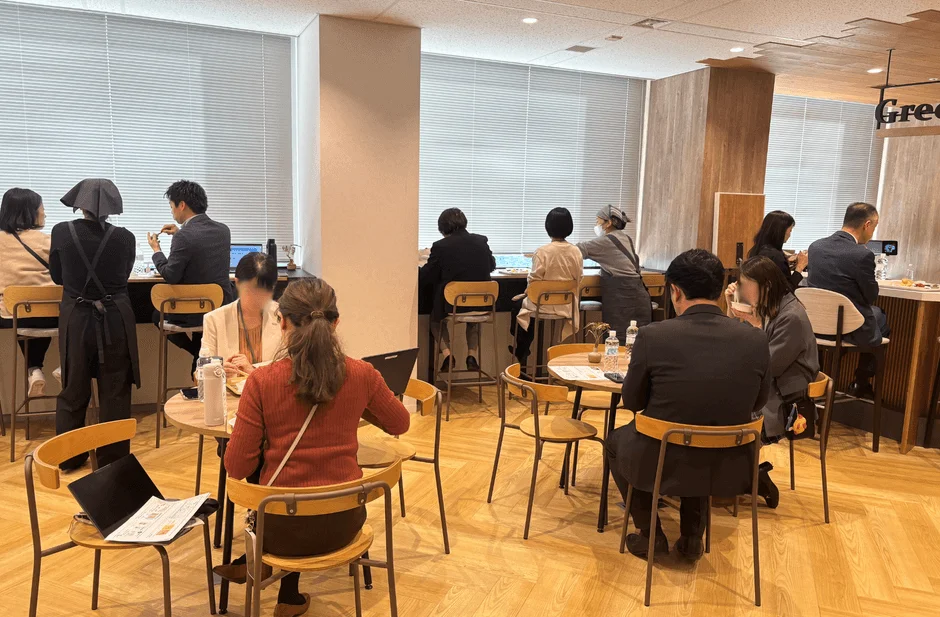 明るくモダンな内装のカフェまたは休憩スペースで、多くの人が食事や休憩を楽しんでいます。カウンター席やテーブル席があり、カジュアルな雰囲気です。