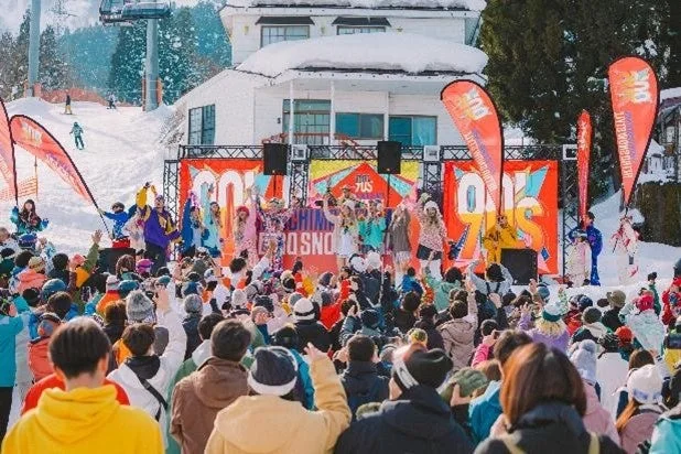 雪山を背景にしたスキー場で開催されている屋外ライブイベントの様子です。ステージにはパフォーマーがおり、多くの観客が音楽を楽しんでいます。90年代をテーマにしたイベントのようです。