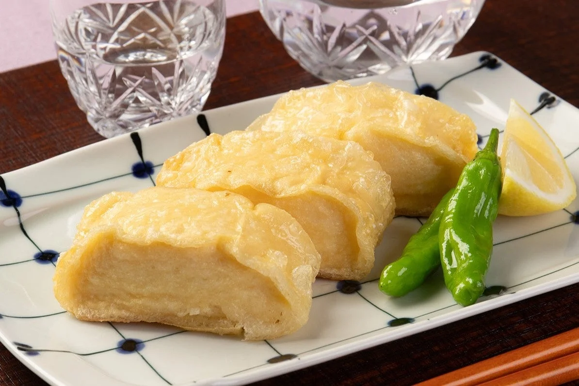 白い皿に盛り付けられた、こんがりと揚げられた厚揚げが3切れ並んでいます。付け合わせに緑色のししとうとレモンが添えられ、背景にはカットグラスと箸が見えます。和食の温かい一品料理の風景です。