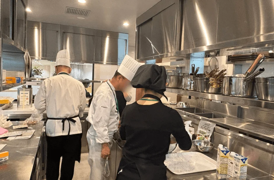 清潔なステンレス製の業務用キッチンで、シェフを含む3人のスタッフが調理作業をしている様子です。食材や調理器具が整然と並べられています。