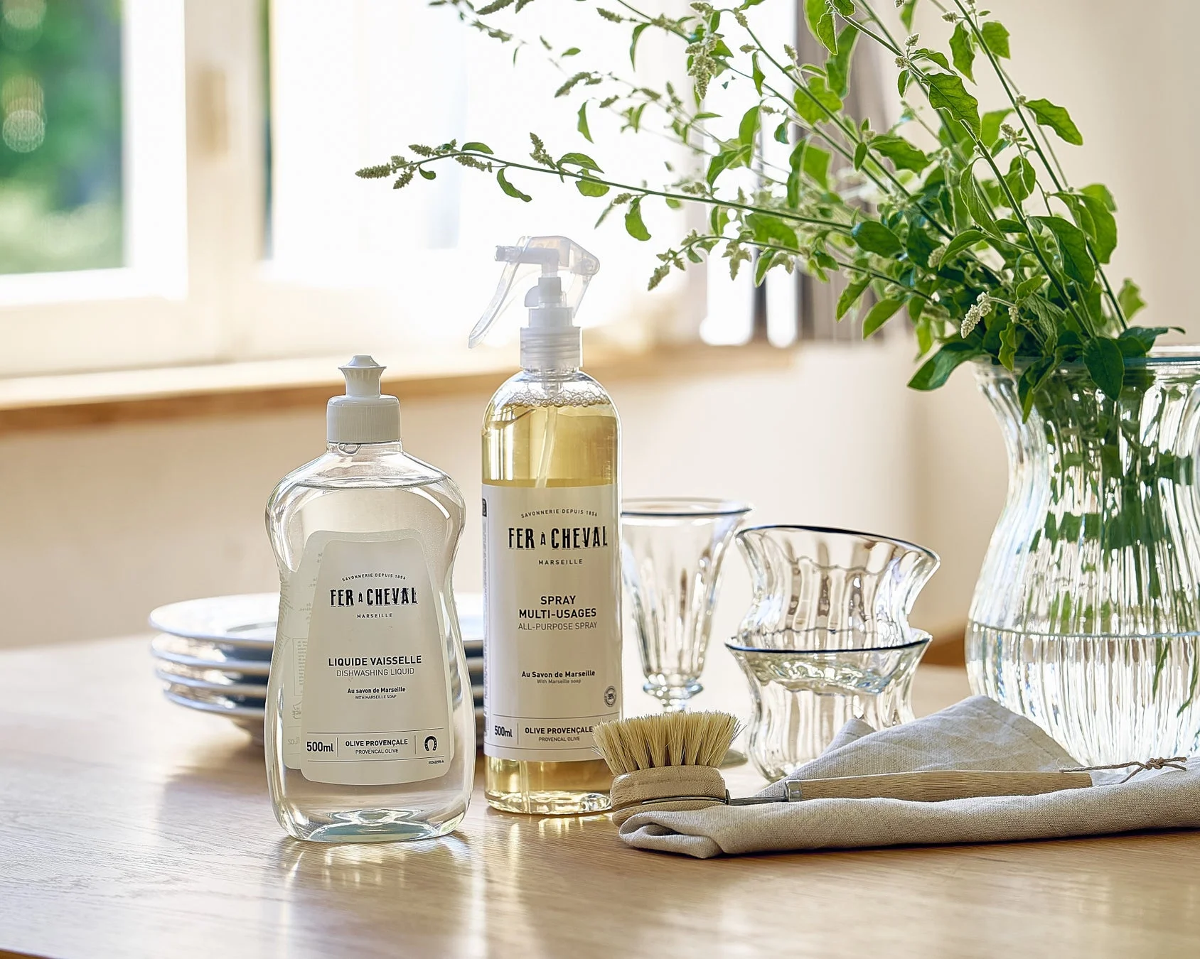 FER+CHEVALの食器用洗剤と多目的スプレー、木製ブラシ、ガラスの器、植物が並べられた清潔感あふれるキッチンの風景