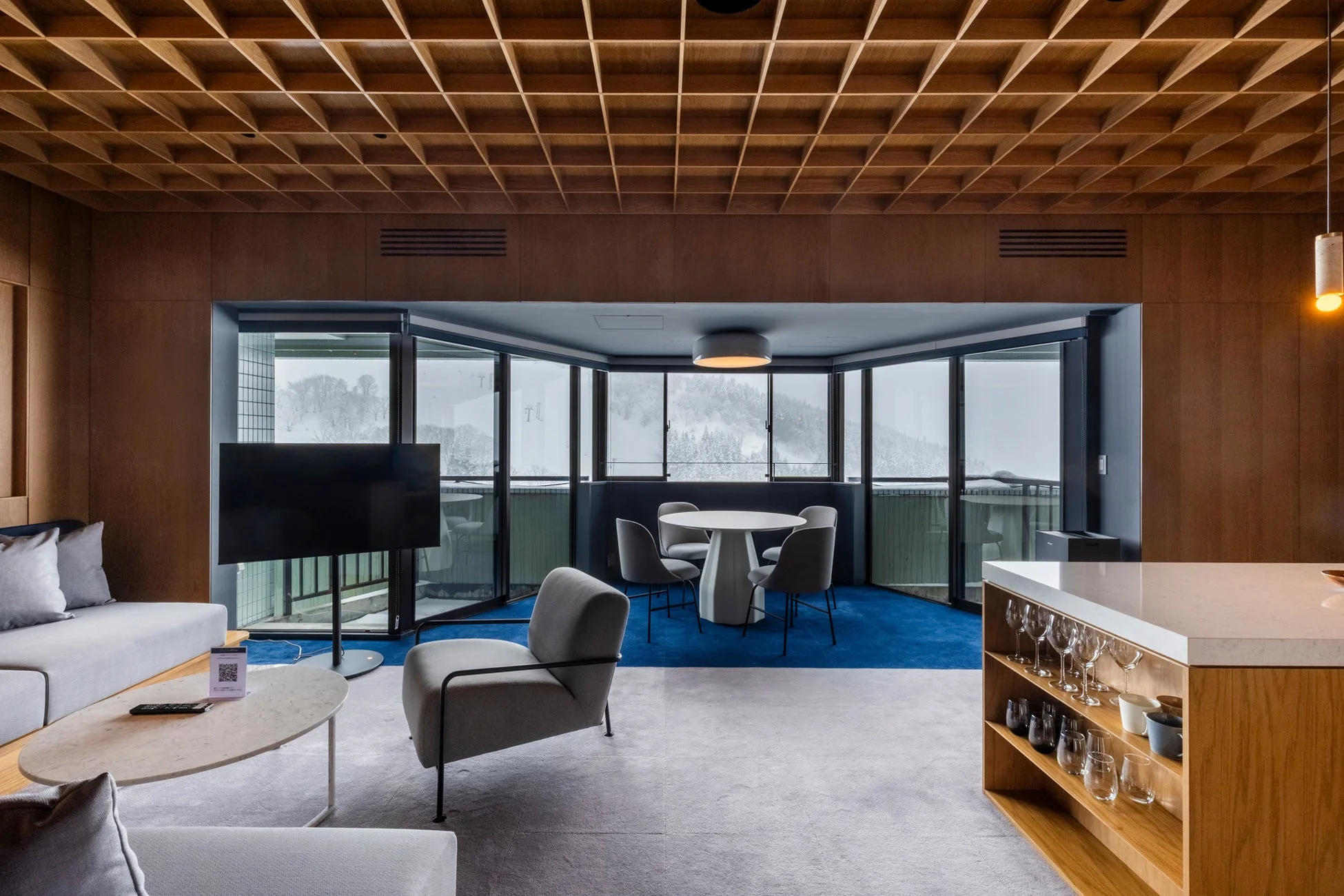 モダンなリビングダイニング空間で、木製格子の天井と雪山の景色が特徴的です。ソファ、ダイニングセット、テレビ、カウンターが配され、洗練されたリゾートホテルの雰囲気を醸し出しています。