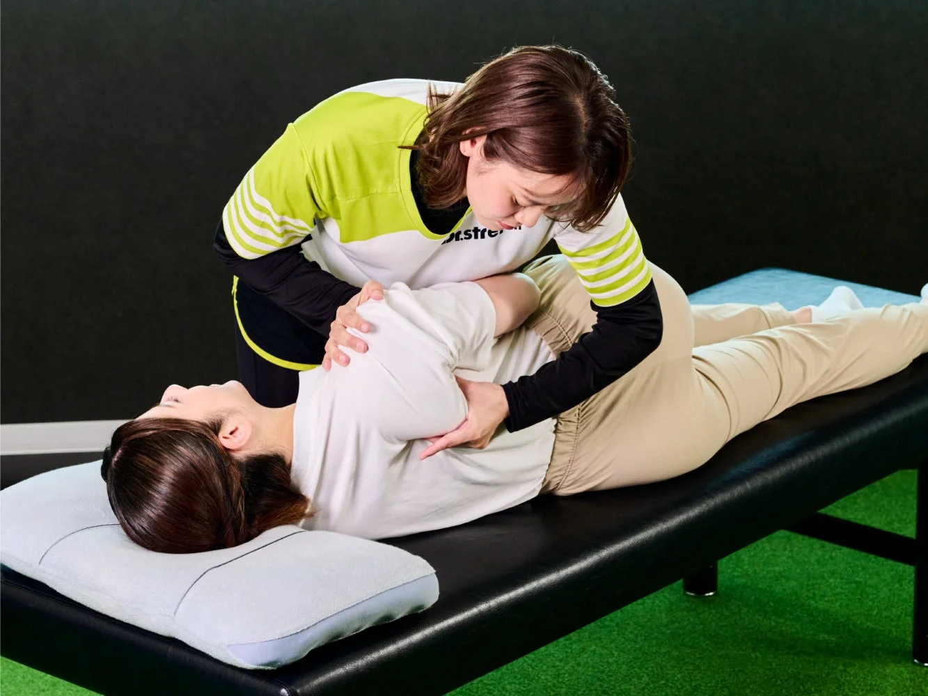 女性のトレーナーが、ベッドに横たわる女性の背中や肩をストレッチしている様子