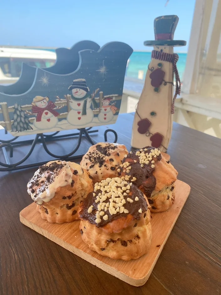 青い海と空を背景に、チョコレートなどで飾られた4つのシュークリームが木製ボードに並んでいます。