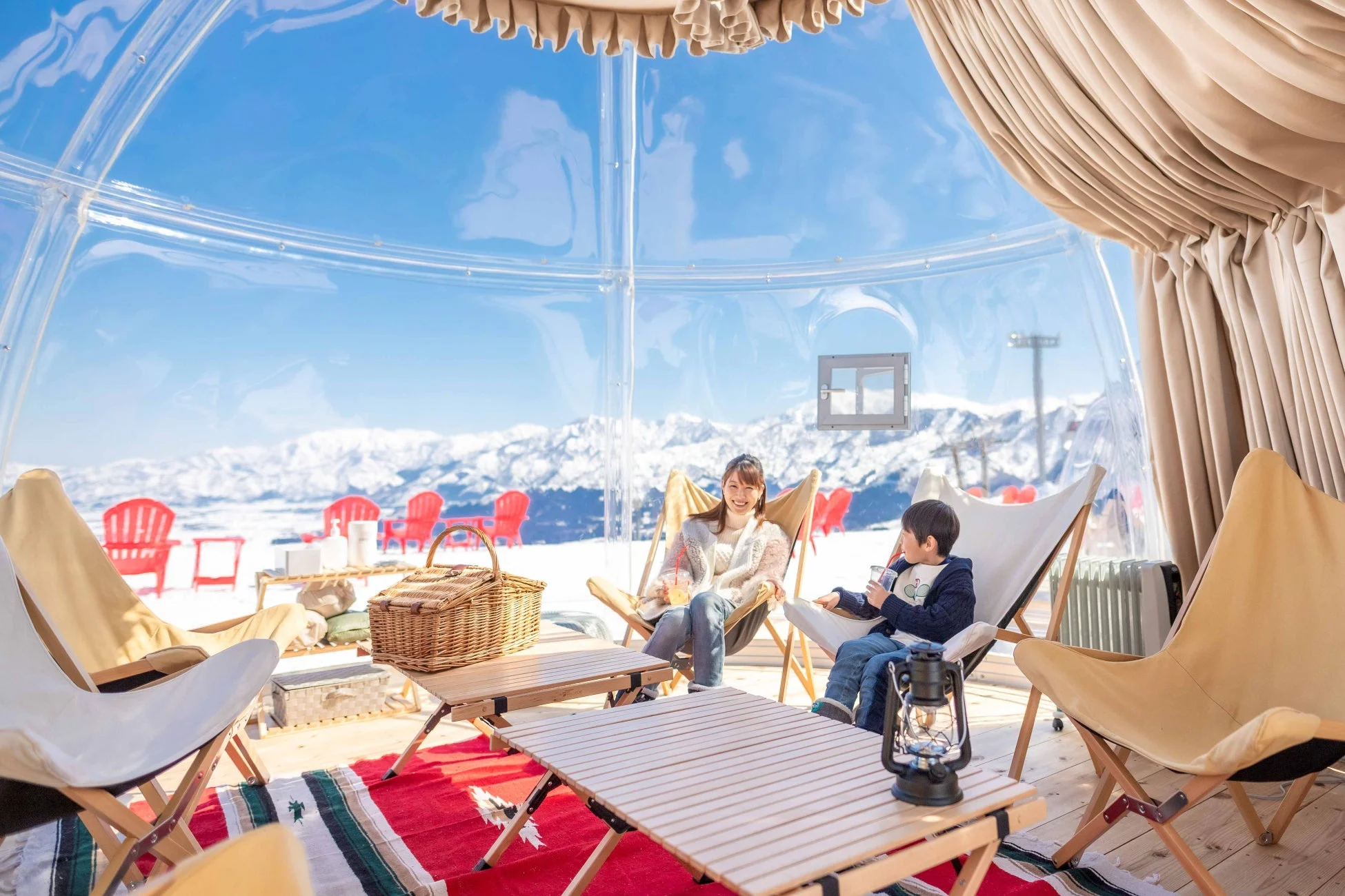 透明なドームテントの中で、女性と子供が雪景色を眺めながらリラックスしている様子。冬のグランピング体験を楽しんでいる光景です。