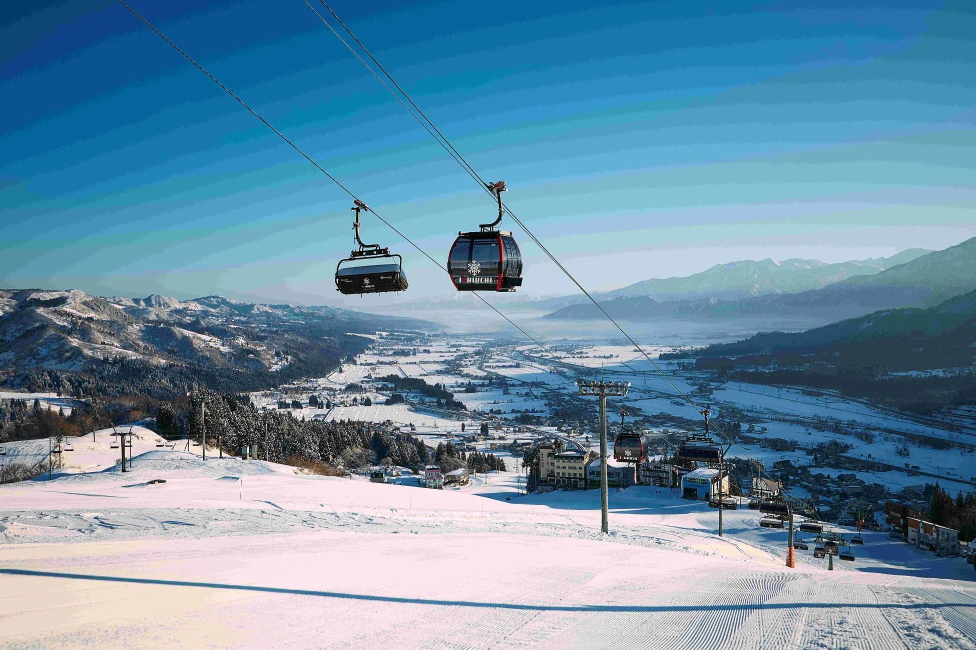 雪に覆われた山々と広大なゲレンデが広がるスキー場の風景です。青空の下、ゴンドラとチェアリフトが運行し、遠くには雪化粧した街並みが見渡せます。冬の美しい景色が印象的です。