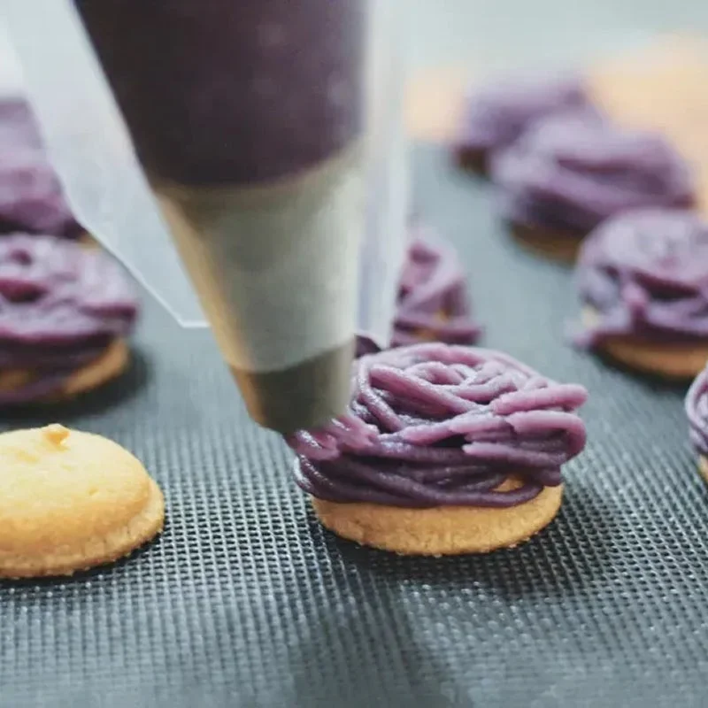 絞り袋から紫色のクリームが小さな焼き菓子の上に絞り出され、モンブランのようなデザートが作られている様子を捉えたクローズアップ。