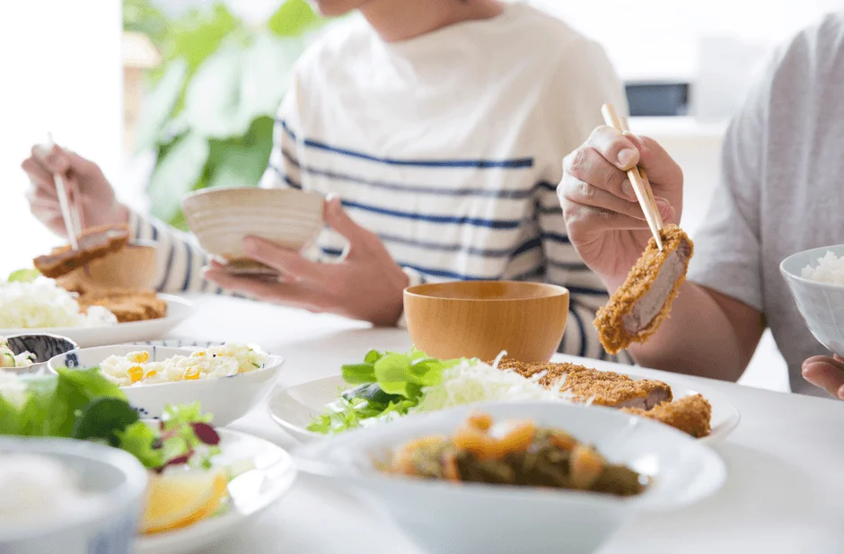二人の人物が食卓を囲み、箸を使って食事をしている様子です。メイン料理はとんかつで、ご飯やサラダなどの副菜も並べられた和食の定食を楽しく味わっている場面が捉えられています。
