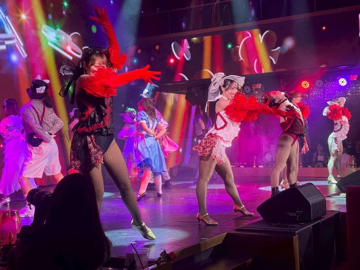 アリス風の衣装を身につけたダンサーたちがステージで活気あふれるパフォーマンス