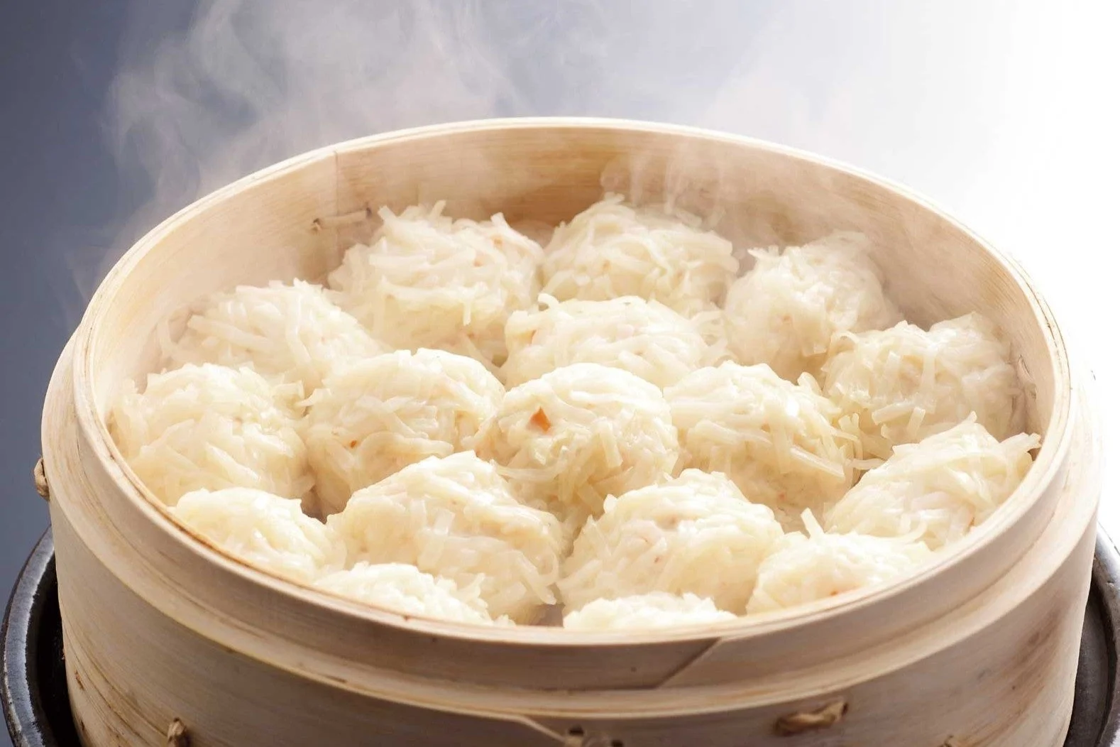 竹製の蒸籠に盛られた、湯気が立ち上る熱々のシュウマイのアップです。白いシュウマイの表面には細かく刻まれた具材がまぶされており、出来立ての美味しそうな雰囲気を醸し出しています。