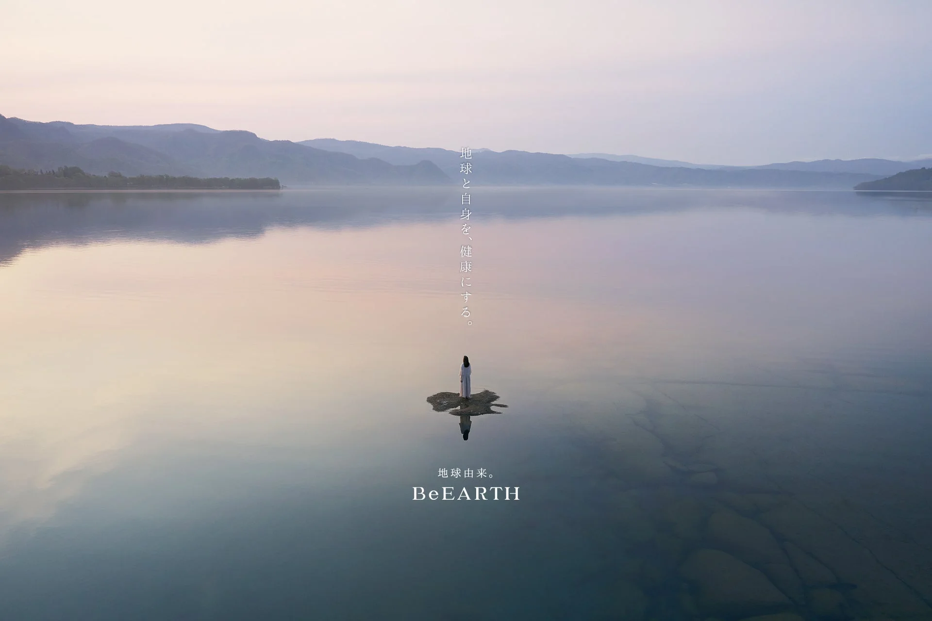広大な湖の中央に立つ一人の人物が、水面に映る空と山々を背景に描かれています。静寂に包まれた幻想的な風景で、「地球と自身を、健康にする。」というメッセージが添えられています。