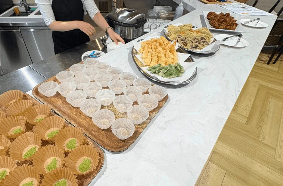 ケータリングかパーティー会場で、ビュッフェ形式の食事が並べられています。海老や野菜の天ぷら、唐揚げ、そしてカップに入ったデザートなど、多彩な料理が用意されており、配膳の準備がされている様子です。