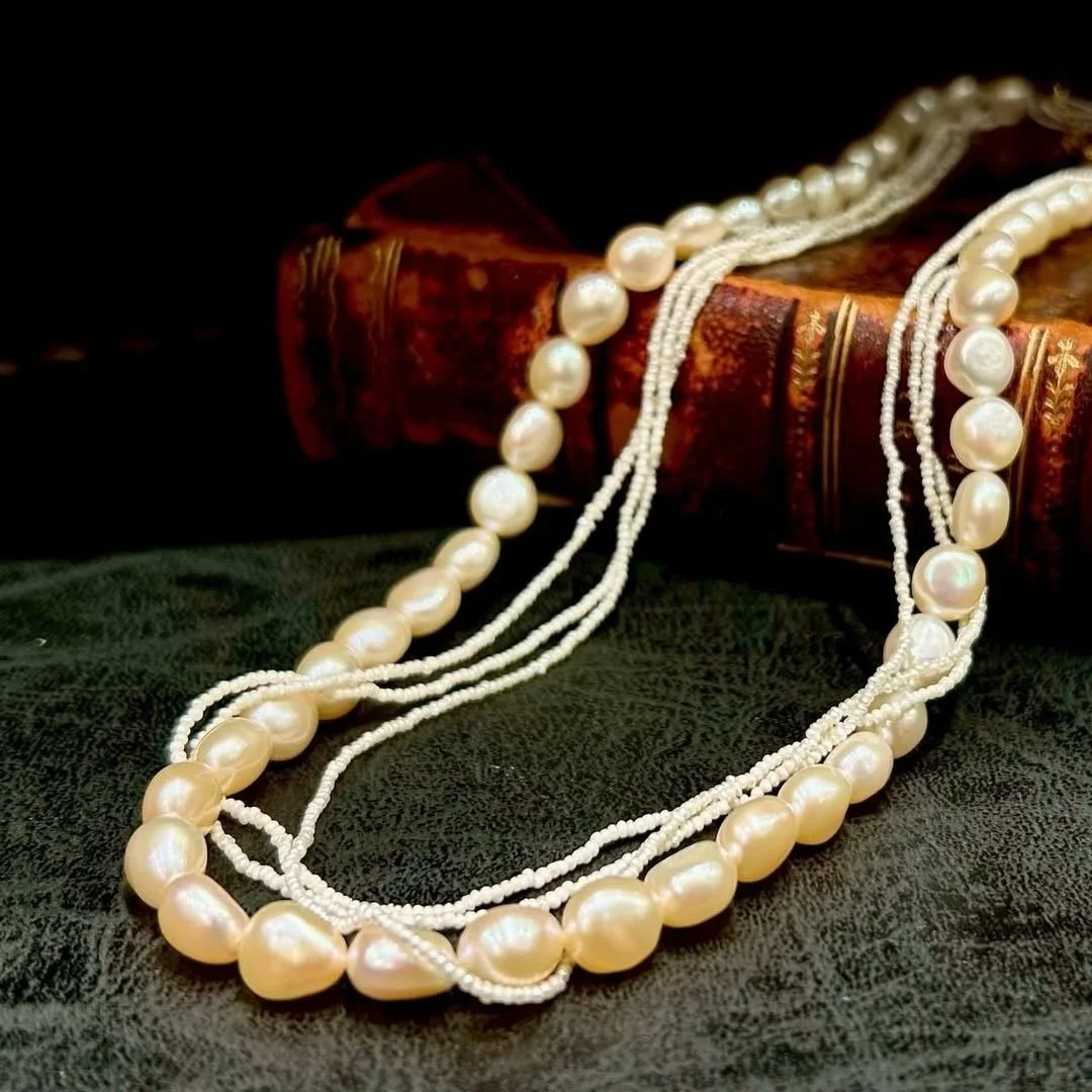 クリーム色のバロックパールと小粒ビーズが何連にも重なったネックレスが、アンティーク調の革製らしき本のそばに置かれている様子