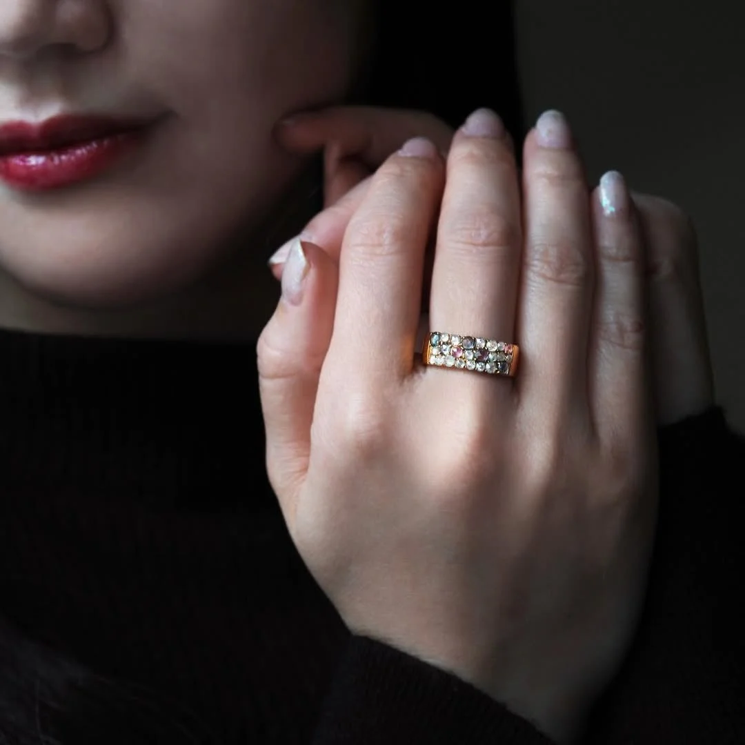 黒い服を着た女性の手が写っており、薬指にはカラフルな宝石が散りばめられたゴールドの指輪が輝いています
