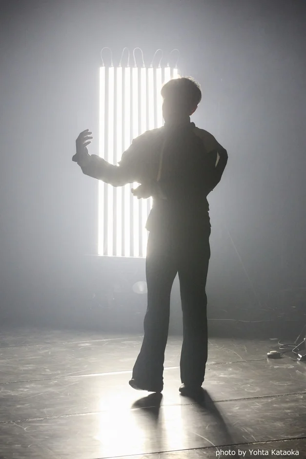 明るい垂直の光を背景に、人物がドラマチックにシルエットで表現されたステージパフォーマンスの様子。芸術的で神秘的な雰囲気が漂う。