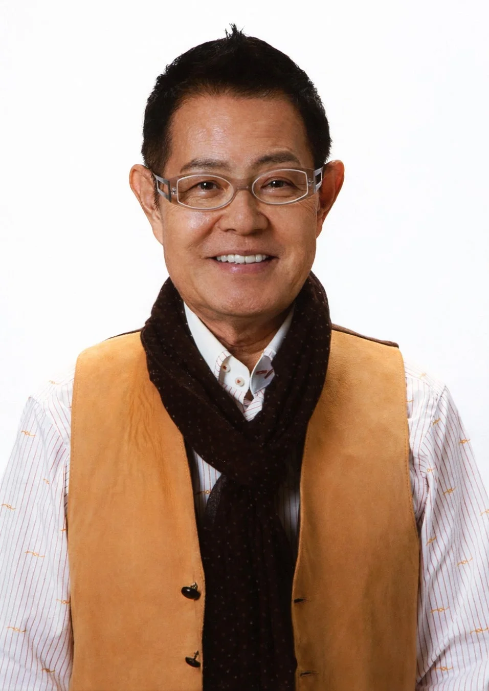 眼鏡をかけ、ストライプのシャツに茶色のスカーフとベストを着用した中高年のアジア人男性が、カメラに向かって笑顔を見せている上半身のポートレート写真