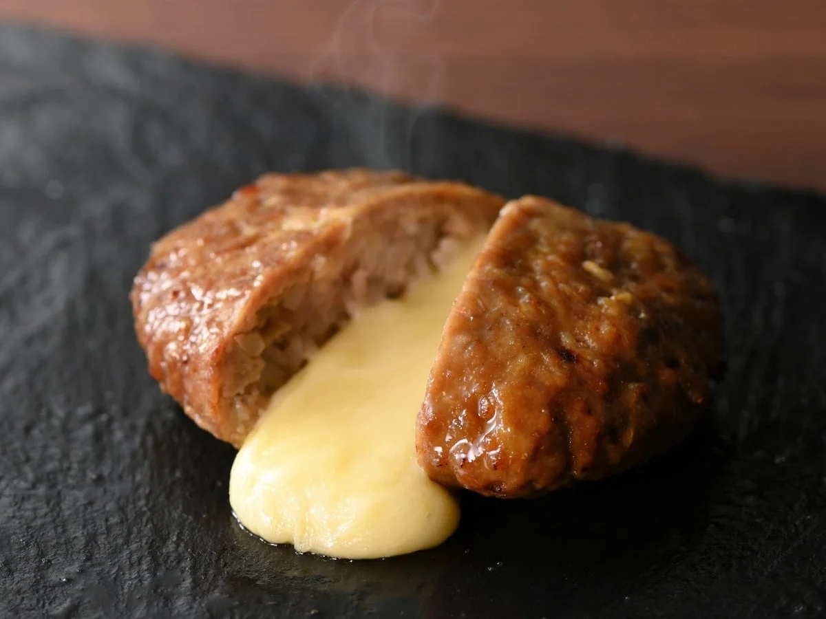 黒い皿に盛られた熱々のハンバーグが写っています。ハンバーグは中央で切り分けられ、中からとろけるチーズがたっぷりあふれ出ています。湯気が立ち上り、食欲をそそる一品です。