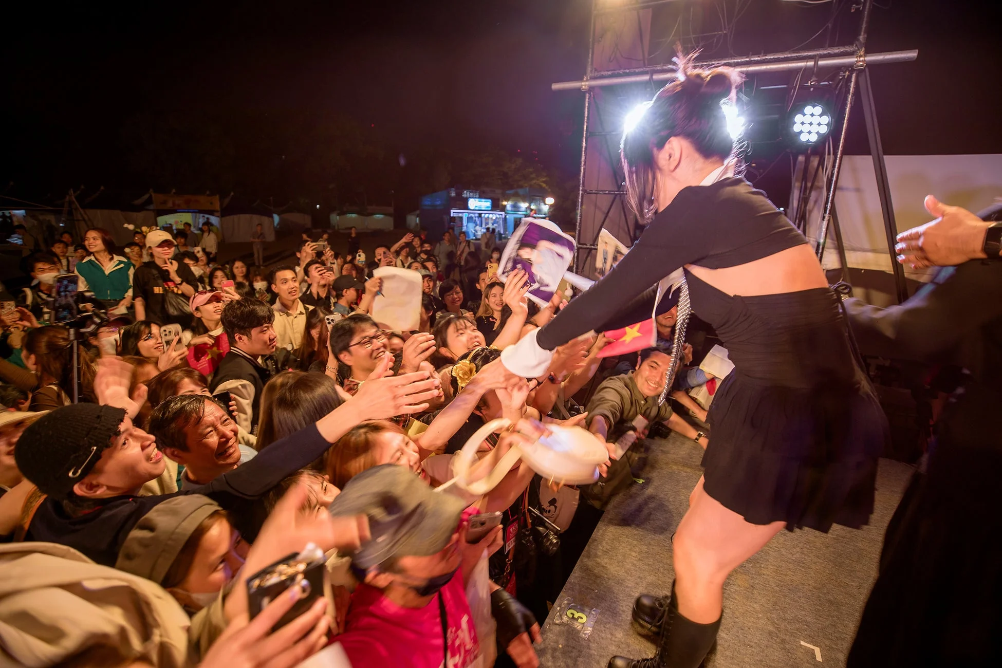 夜の屋外イベントで、ステージ上のパフォーマーが熱狂的な観客と交流している場面