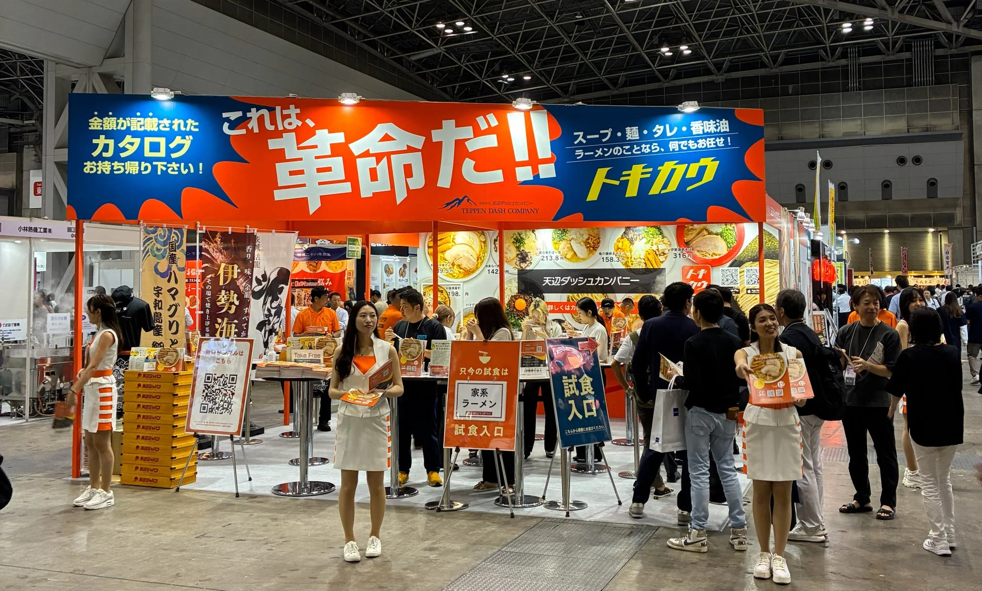 ラーメン関連企業「TEPPEN DASH COMPANY」の展示会ブースで、来場者が試食を楽しんでいる様子が写っています。