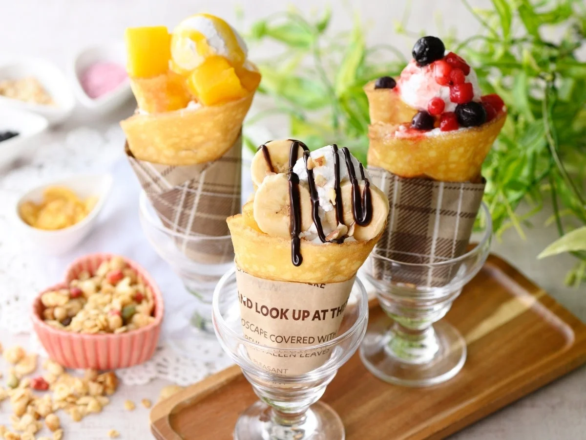 3種類のフルーツクレープがグラスに盛られ、マンゴー、バナナ、ミックスベリーがトッピングされています。グラノーラなども添えられた、彩り豊かで美味しそうなデザートの盛り合わせです。