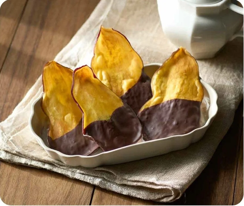 木製のテーブルの上に置かれた白い皿に、チョコレートを半分だけディップしたさつまいものお菓子が盛り付けられています