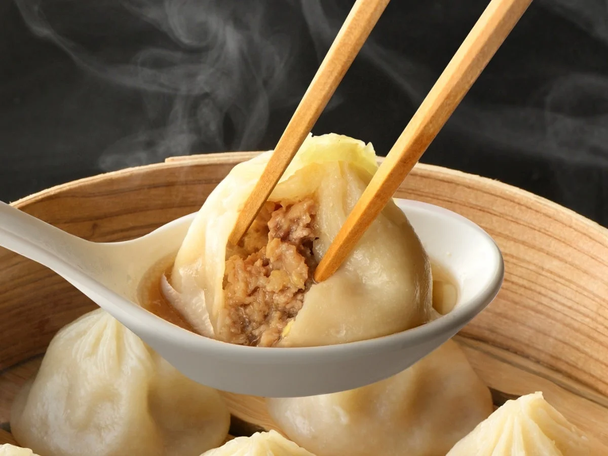熱々の小籠包を箸で割り、肉汁があふれ出る瞬間を捉えたクローズアップ写真。白いスプーンに乗せられ、湯気が立ち上る蒸篭の中の他の小籠包も見える。食欲をそそる一枚。