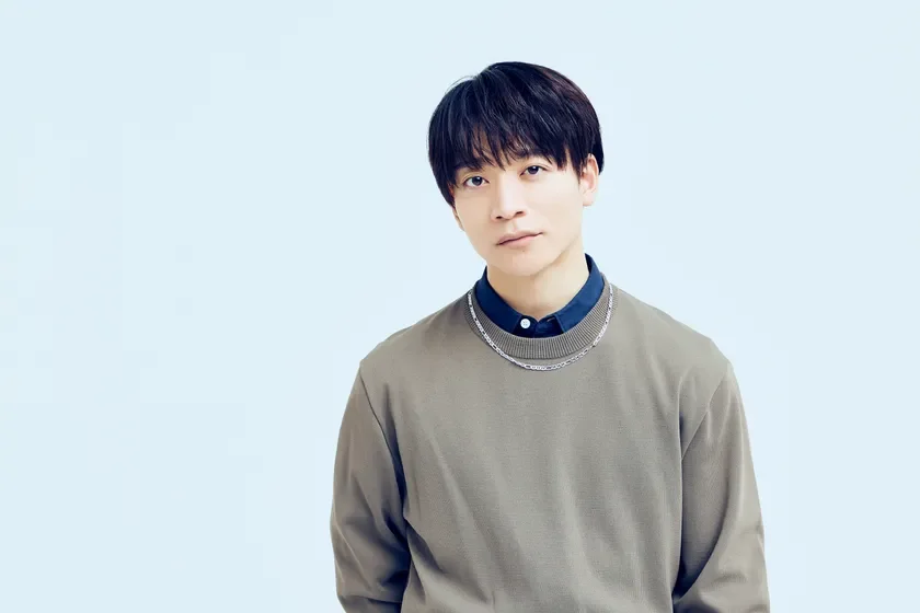 若い日本人男性が、グレーのセーターと紺色のシャツを着て、カメラを見つめているポートレート写真です。