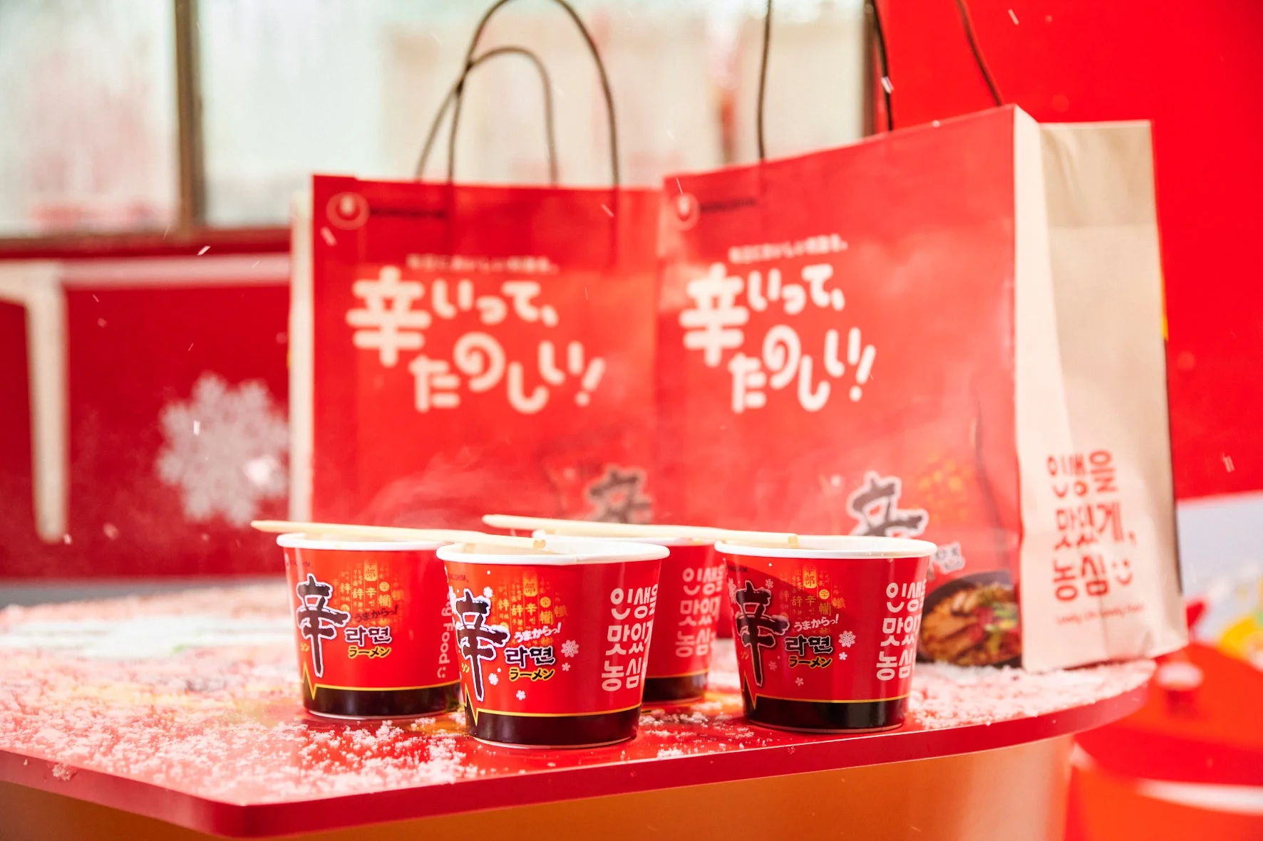 赤い背景に「辛ラーメン」のカップ麺が複数並べられ、奥には「辛いって、たのしい！」と書かれた赤いショッピングバッグが見える。