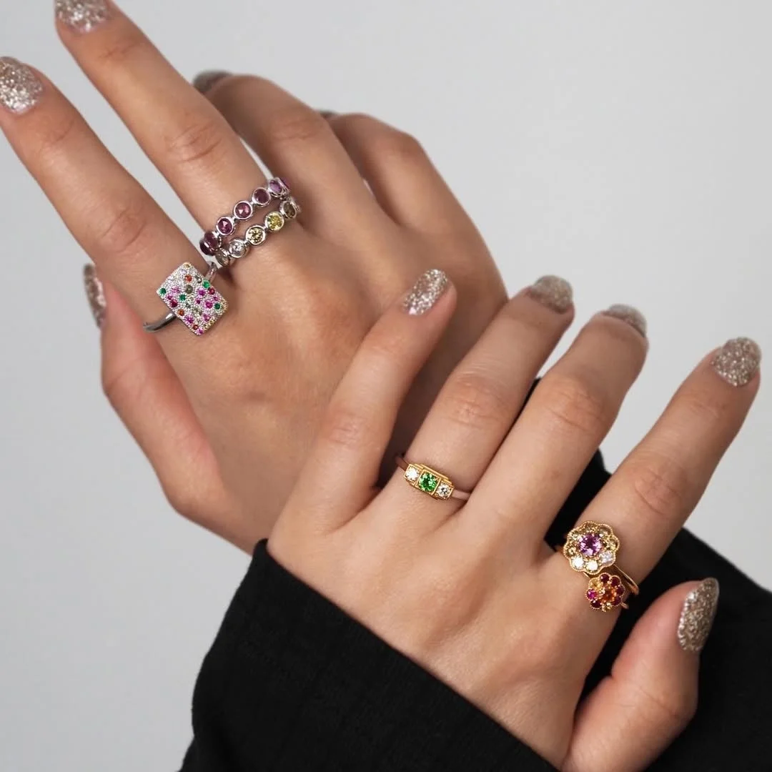 グリッターネイルを施した女性の両手が、カラフルな宝石があしらわれた複数の指輪を着用している様子