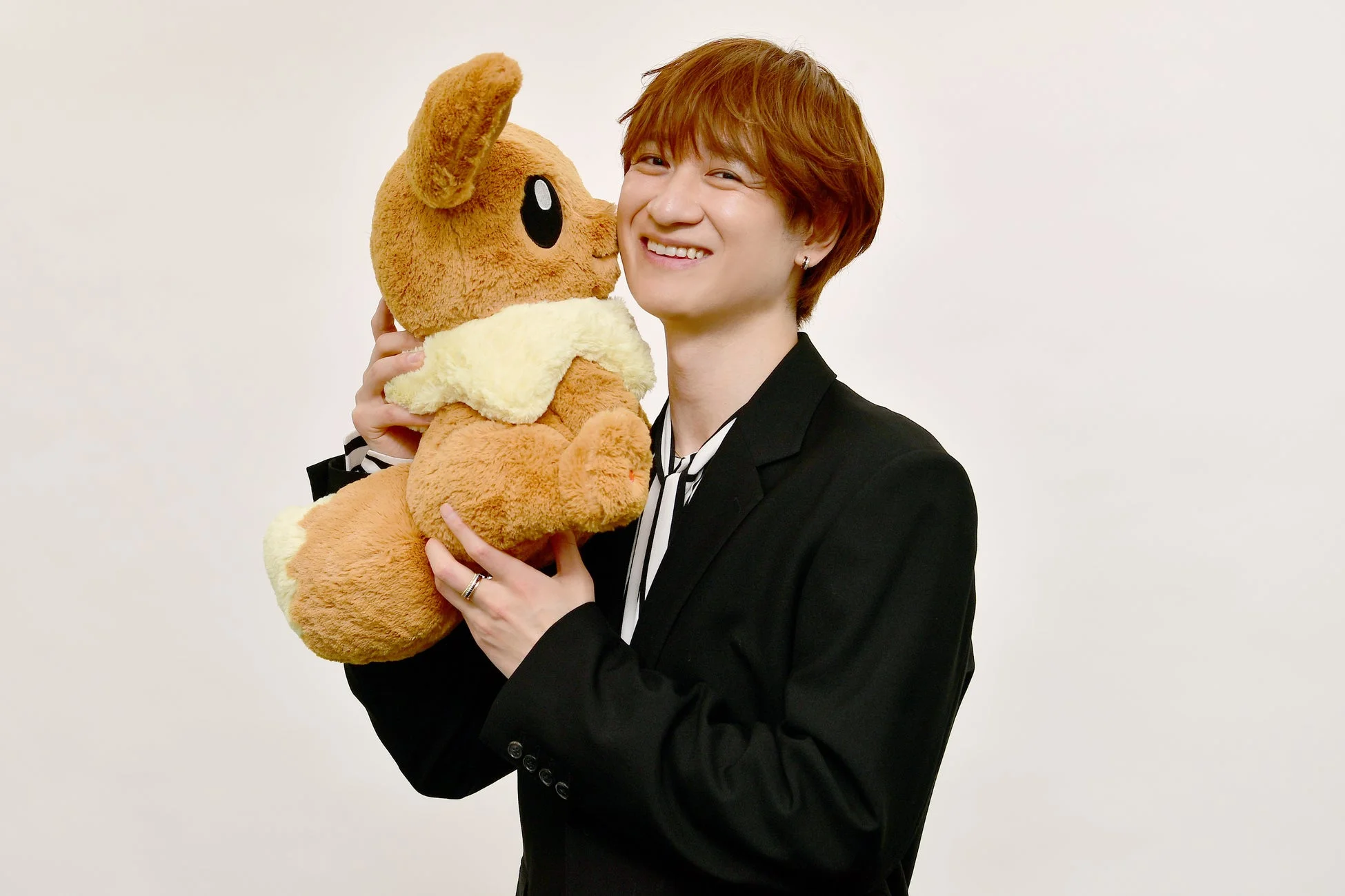 茶色の髪の若い男性が、大きなイーブイのぬいぐるみを抱え、満面の笑みを浮かべている。黒いジャケットとストライプのシャツを身につけ、白い背景の前で親しみやすい表情を見せている。