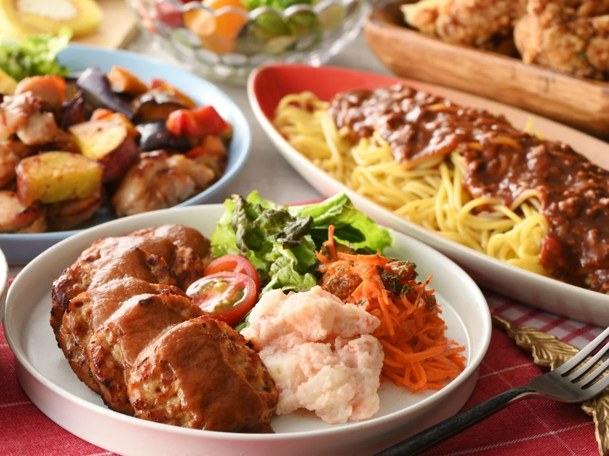 ハンバーグやミートソーススパゲッティ、ポテトサラダなどが並べられた、彩り豊かな洋食の食卓風景です。ボリューム満点で美味しそうな家庭料理が複数皿に盛り付けられています。
