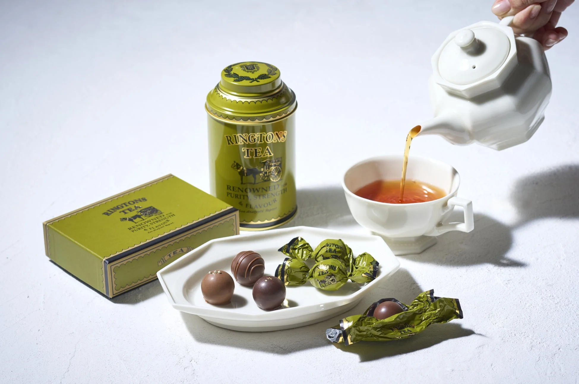 リントンズの紅茶缶と箱、そしてチョコレートが並べられたテーブルで、白いティーポットからカップに紅茶が注がれている様子