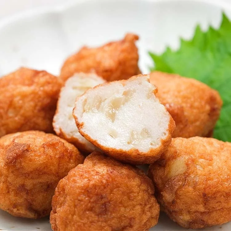 揚げたてのさつま揚げが複数個、白い皿に盛り付けられています。手前の一つは半分に切られ、白い魚肉と具材が確認できます。奥には緑色の葉が添えられており、食欲をそそる日本の惣菜です。