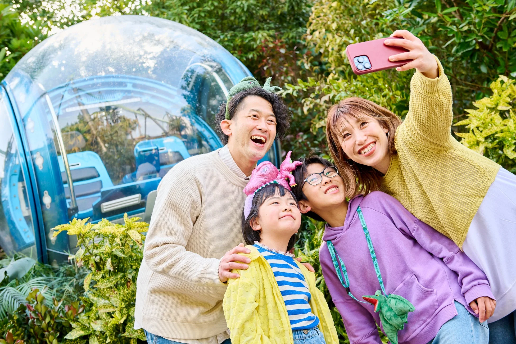 ジュラシックワールドのようなテーマパークで、恐竜のアクセサリーを身につけた家族４人が屋外で楽しそうにセルフィーを撮っている