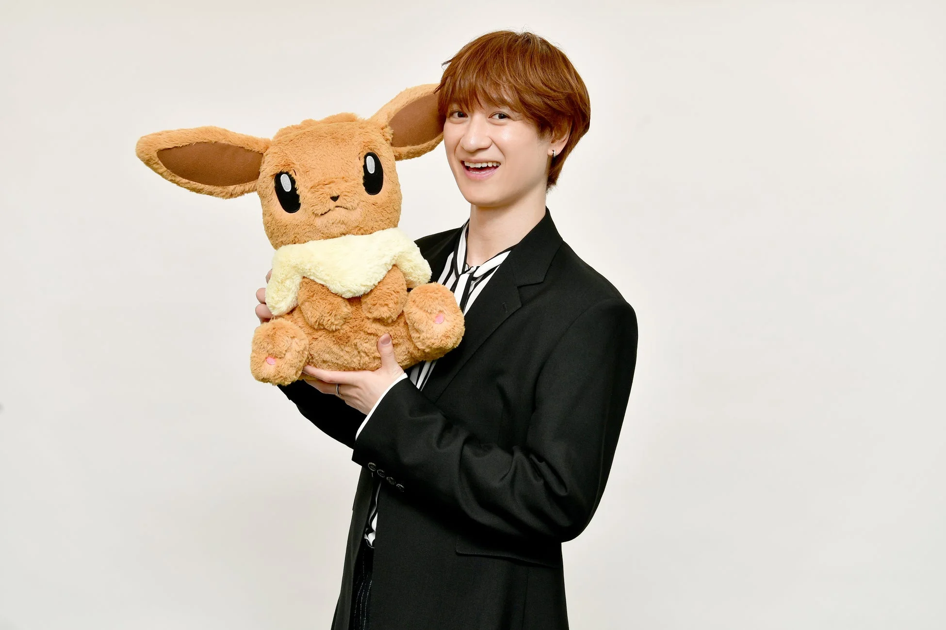 茶髪の若い男性が、黒いジャケットとストライプのシャツを着用し、大きなイーブイのぬいぐるみを抱えて笑顔を見せている。背景は白一色で、明るく親しみやすい雰囲気のポートレート写真です。
