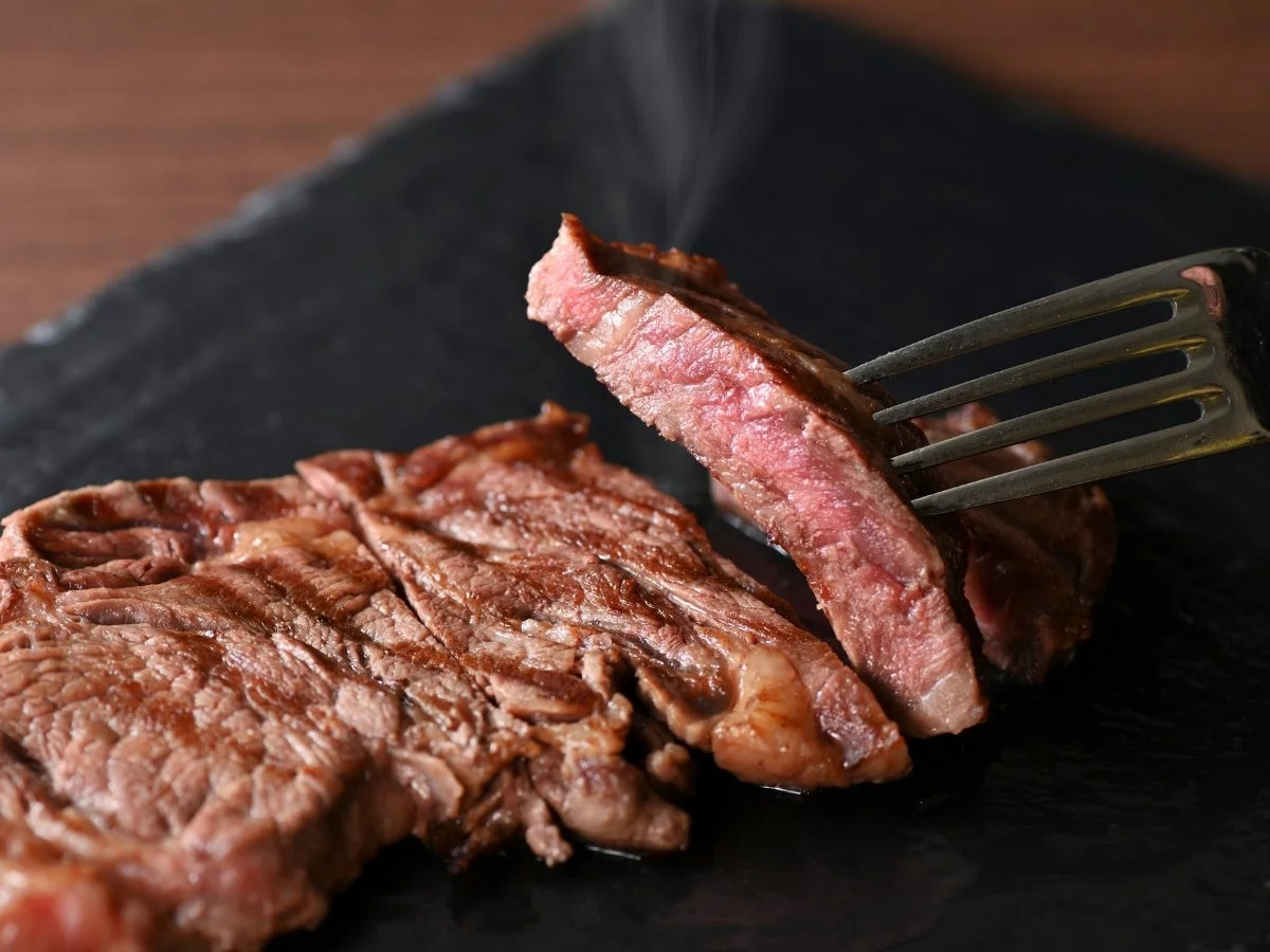 焼きたてのステーキが黒い皿に盛り付けられ、一切れがフォークで持ち上げられている様子です。肉は美しいミディアムレアに焼かれており、湯気が立ち上っています。食欲をそそる一品です。