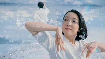 白い服の女性が水しぶきの中で優雅なポーズをとり、背景には白い服の男性が立つ